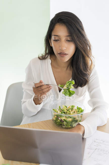Портрет молодого архітектора, який їсть змішаний салат за столом, дивлячись на ноутбук. — стокове фото