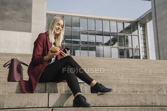 Femme d'affaires blonde déjeunant dehors et utilisant un smartphone — Photo de stock