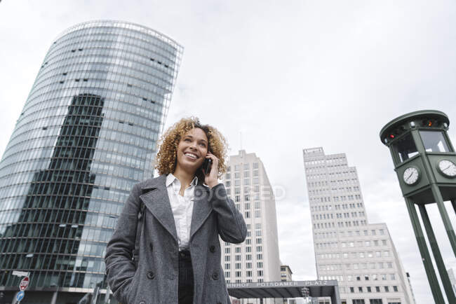 Mujer sonriente al teléfono con edificios de oficinas en segundo plano, Berlín, Alemania - foto de stock