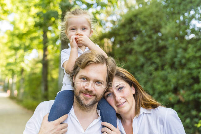 Portait of happy family in a park with father carrying little daughter on his shoulders — Stock Photo
