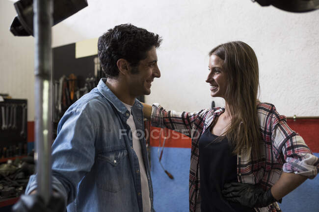 Vista lateral de la joven mujer sonriente y el hombre mirándose mientras están de pie en el servicio de reparación de automóviles y la comunicación - foto de stock