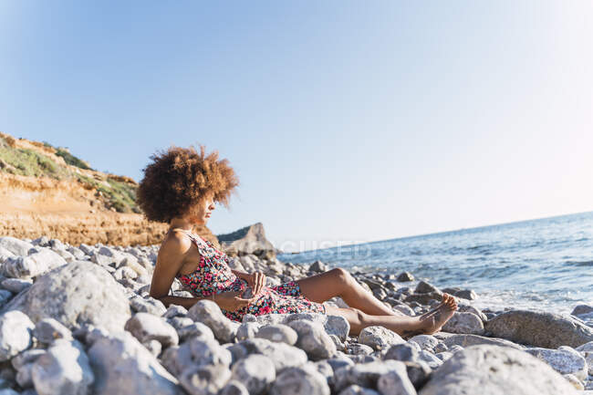 Giovane donna che indossa abito estivo e seduto sulla spiaggia di pietra — Foto stock