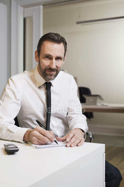 Portrait of confident businessman sitting at desk in office taking notes — Stock Photo