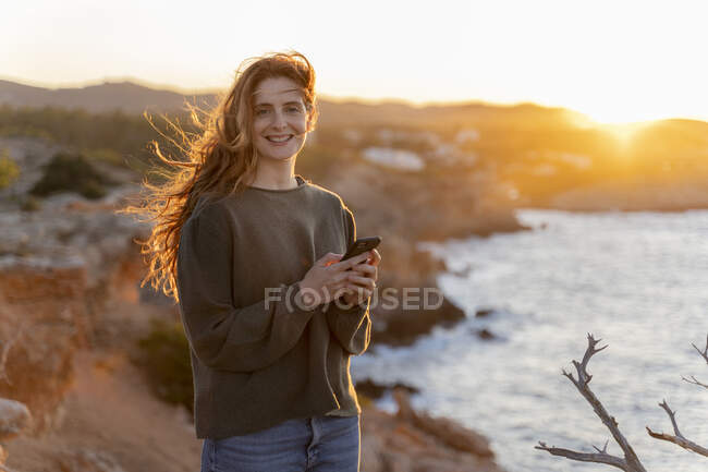 Ritratto di giovane donna rossa sorridente con cellulare sulla costa al tramonto, Ibiza, Spagna — Foto stock
