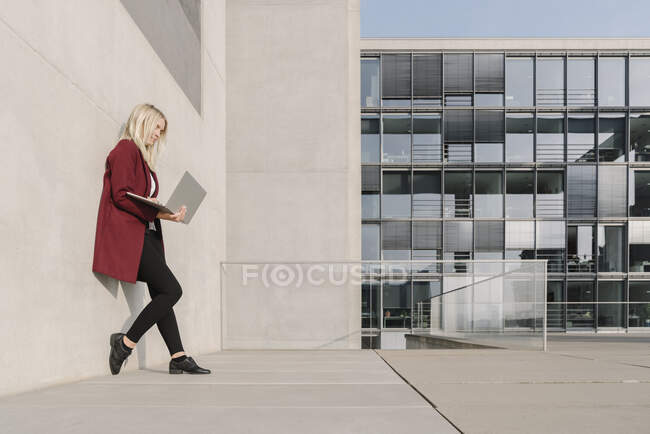Femme d'affaires blonde utilisant un ordinateur portable et s'appuyant sur un mur — Photo de stock