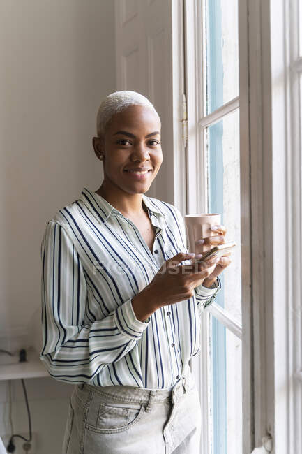 Retrato de mujer sonriente con teléfono celular y taza de café en la ventana - foto de stock