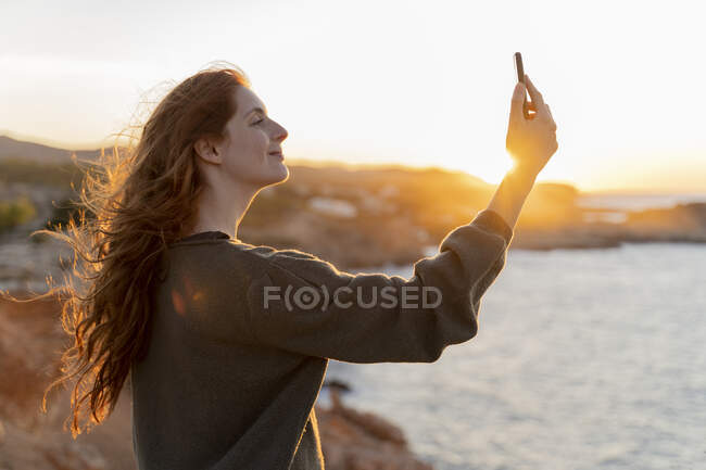 Giovane donna dai capelli rossi che si fa un selfie sulla costa al tramonto, Ibiza, Spagna — Foto stock