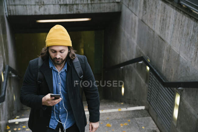 Hombre con auriculares saliendo de la estación de tren en el día lluvioso mirando el teléfono celular - foto de stock