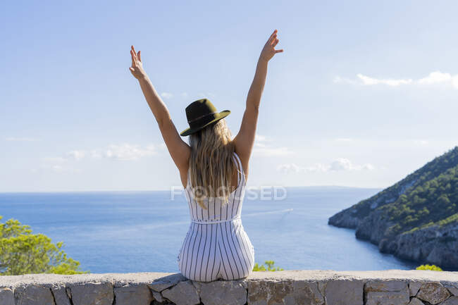 Young woman sitting on a wall with raised arms and looking at distance, viewpoint in Ibiza — Stock Photo