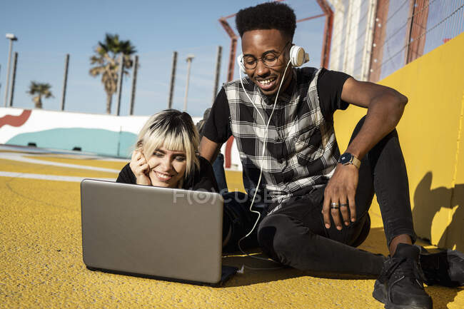 Retrato de pareja joven sonriente usando auriculares y computadora portátil al aire libre - foto de stock