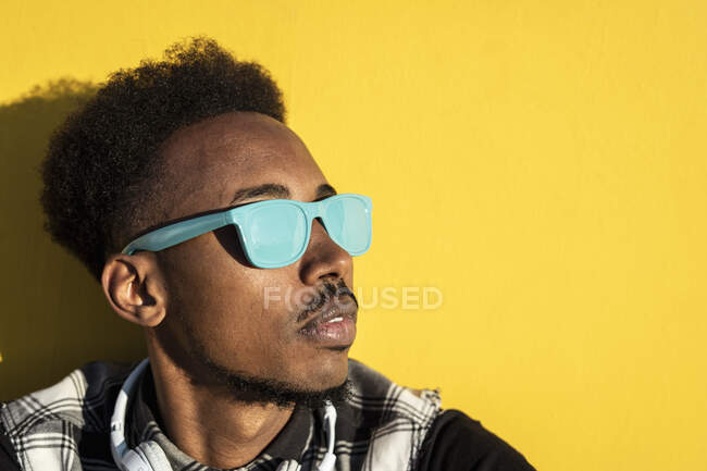 Portrait de jeune homme portant des lunettes de soleil bleu clair contre un mur jaune — Photo de stock