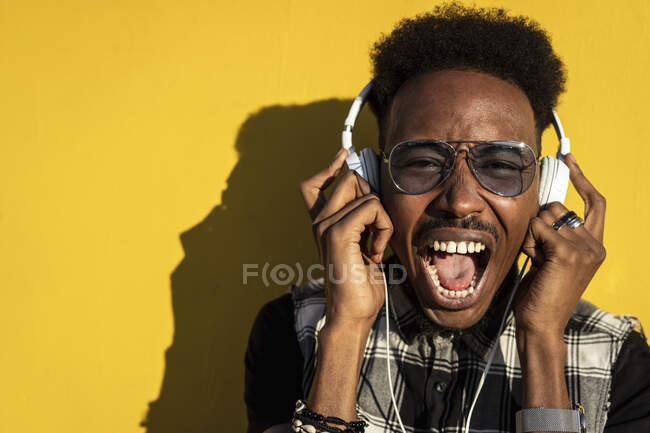 Retrato de un joven gritando contra una pared amarilla escuchando su canción favorita con auriculares - foto de stock