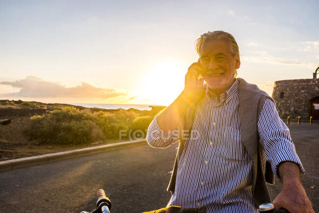 Uomo anziano che utilizza smartphone in bicicletta al tramonto, Tenerife — Foto stock