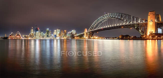 Foto panoramica del ponte illuminato Sydney Harbor Bridge sul fiume contro il cielo di notte, Sydney, Australia — Foto stock
