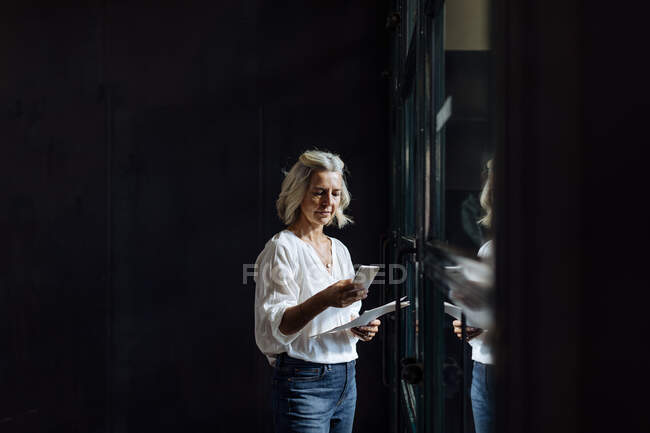 Casual donna d'affari matura utilizzando smartphone alla finestra in ufficio loft — Foto stock