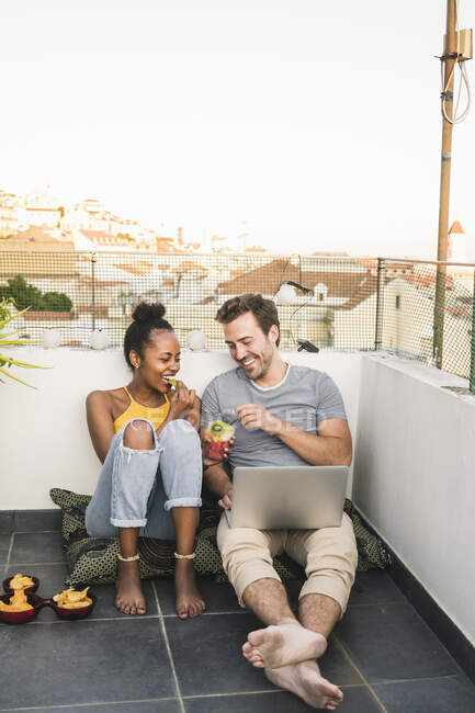 Glückliches junges Paar mit Laptop sitzt abends beim Imbiss auf dem Dach — Stockfoto