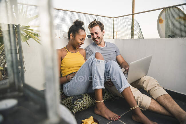 Felice giovane coppia con computer portatile e auricolari seduti sul tetto la sera — Foto stock
