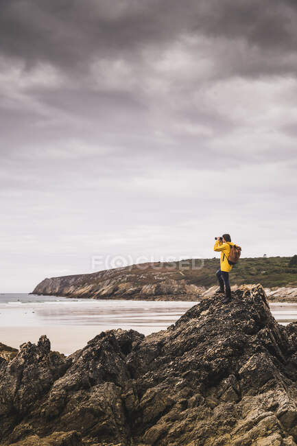 Mujer joven con chaqueta de lluvia amarilla en la playa, mirando a través de prismáticos, Bretagne, Francia - foto de stock