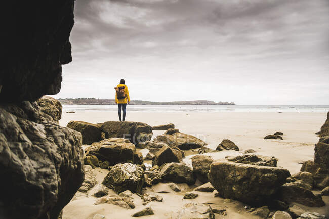 Mujer joven con chaqueta de lluvia amarilla en la playa en la cueva de roca, Bretagne, Francia - foto de stock