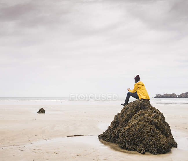 Jeune femme vêtue d'une veste jaune assise sur le rocher à la plage, Bretagne, France — Photo de stock