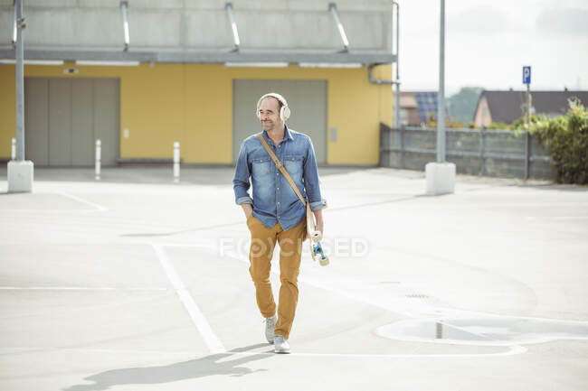 Uomo maturo casual con cuffie e skateboard a piedi sul ponte di parcheggio — Foto stock