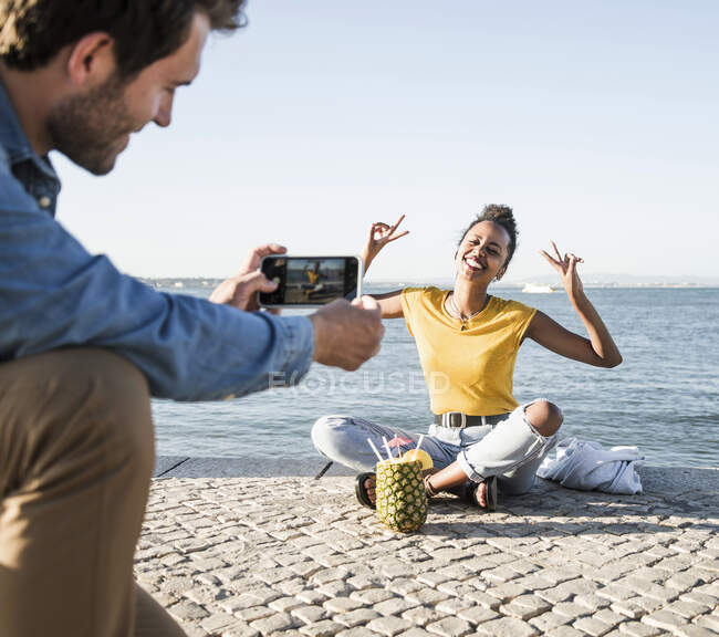 Young man taking a picture of his girlfriend sitting on pier at the waterfront, Lisbon, Portugal — Stock Photo