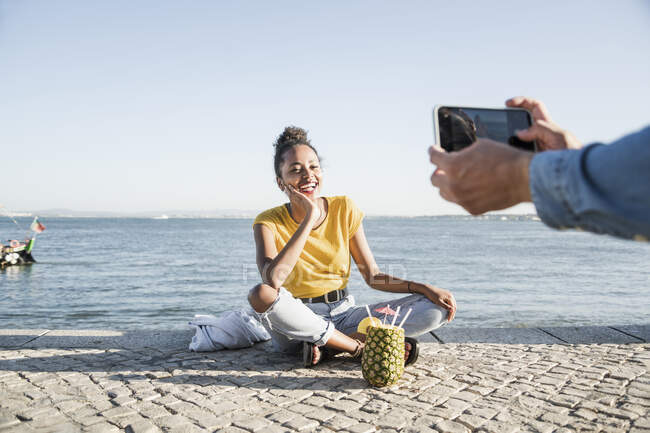 Jeune homme prenant une photo de sa petite amie assise sur un quai au bord de l'eau, Lisbonne, Portugal — Photo de stock