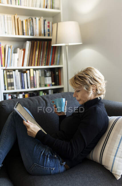 Зрелая женщина отдыхает дома на диване с помощью графического планшета — стоковое фото