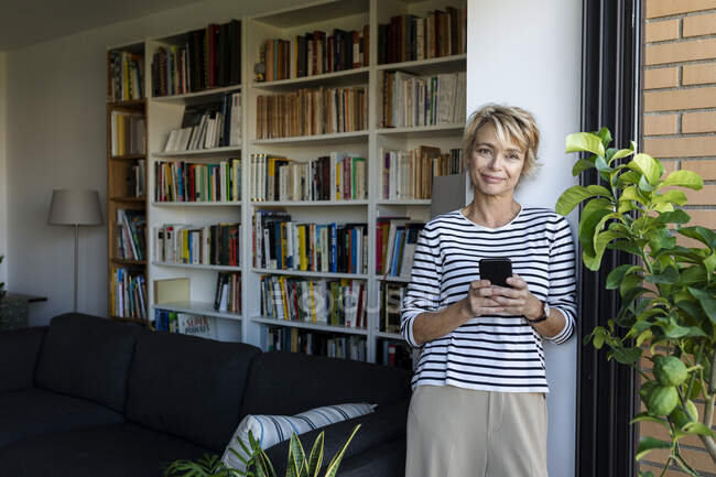 Ältere Frau steht zu Hause am Fenster im Wohnzimmer und hält Smartphone — Stockfoto
