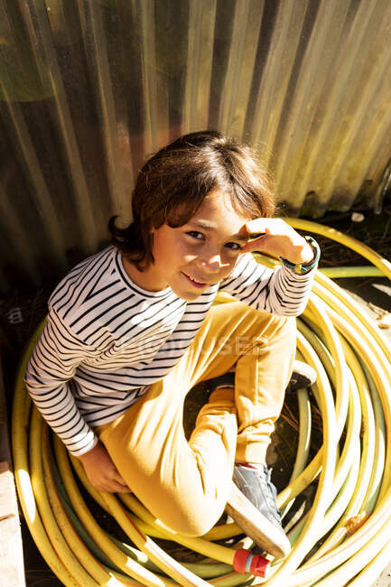 Portrait of smiling boy sitting among rolled up garden hose at plant nursery — Stock Photo