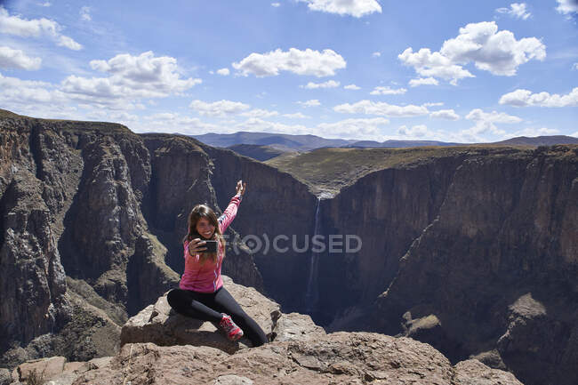 Mujer feliz sentada en la cima de una colina en Maletsunyane Falls tomando una selfie, Lesotho - foto de stock