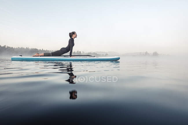Mujer practicando paddle board yoga en el lago Kirchsee por la mañana, Bad Toelz, Baviera, Alemania - foto de stock