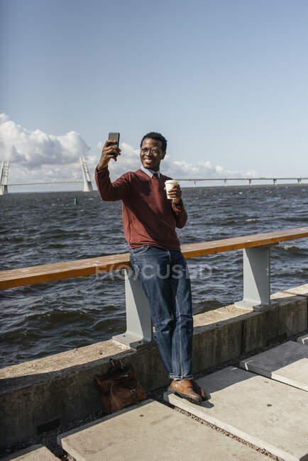 Young man taking smartphone selfie at the sea, holding take out coffee — Stock Photo