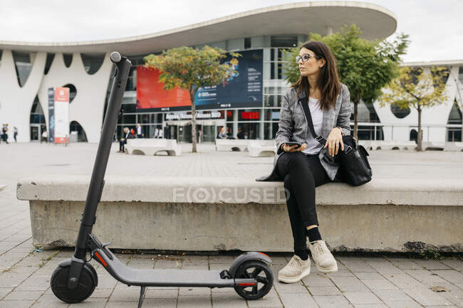 Giovane donna d'affari con smartphone accanto a un e-scooter in città — Foto stock