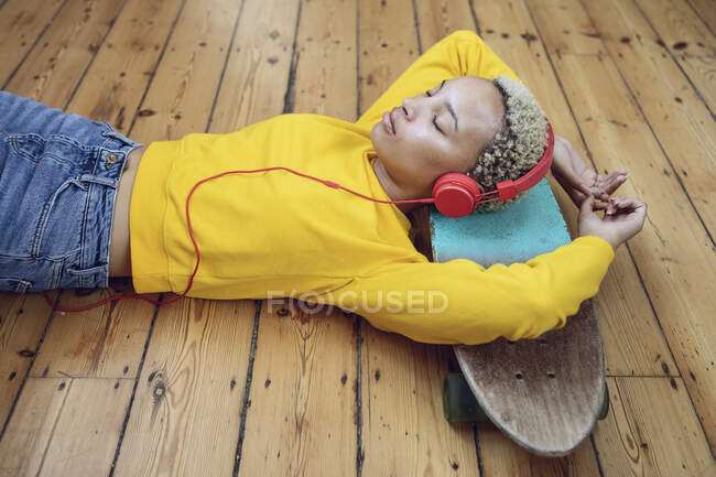 Young woman lying on skateboard on the floor at home listening to music — Stock Photo