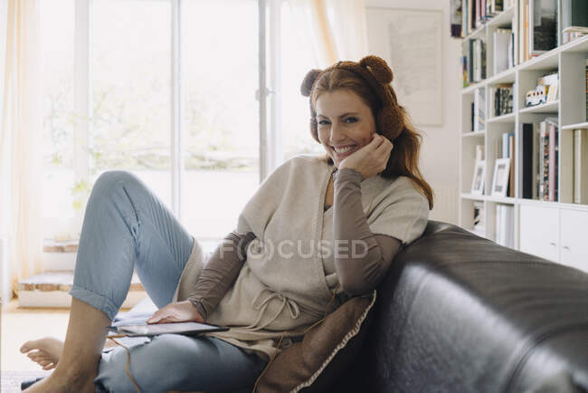 Lächelnde Frau sitzt auf Couch, hört Musik mit digitalem Tablet und Plüsch-Kopfhörer — Stockfoto
