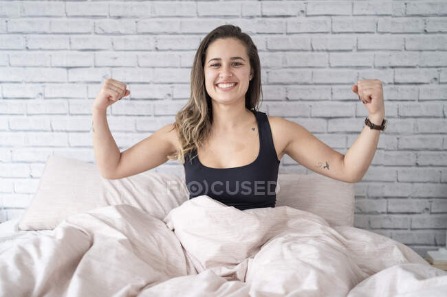 Porträt einer glücklichen jungen Frau, die auf dem Bett sitzt und die Muskeln spielen lässt — Stockfoto