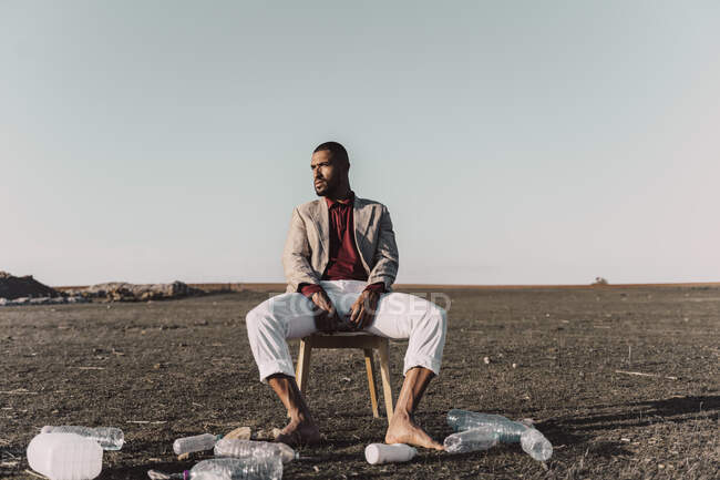 Молодой человек сидит на стуле в окружении пластиковых бутылок в бесплодной земле — стоковое фото