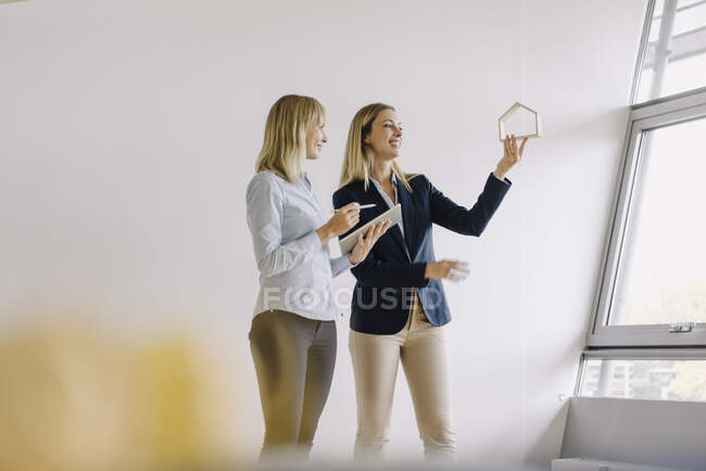 Two young businesswomen with tablet and house model talking in office — Stock Photo