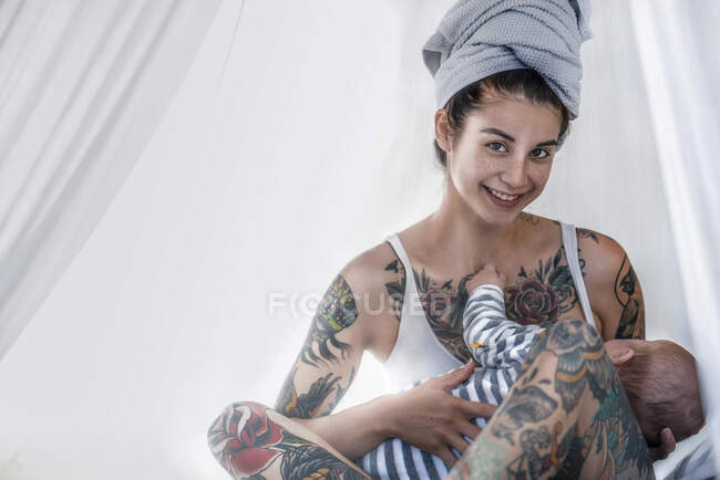 Portrait of smiling tattooed young woman with her baby in canopy bed — Stock Photo