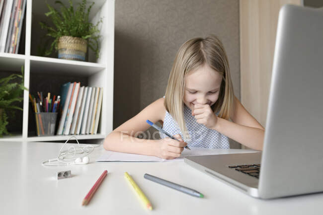 Ragazza ridente seduta a tavola a casa a fare i compiti e utilizzando il computer portatile — Foto stock