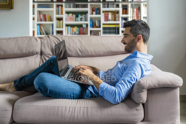 Smiling young man lying on the couch at home using laptop — Stock Photo