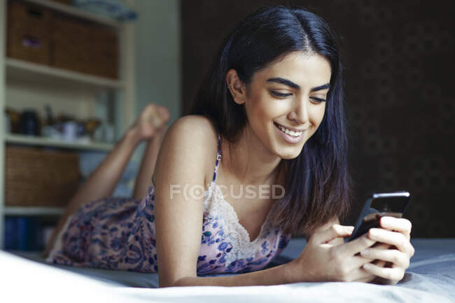 Ritratto di giovane donna sorridente sdraiata sul letto guardando lo smartphone — Foto stock