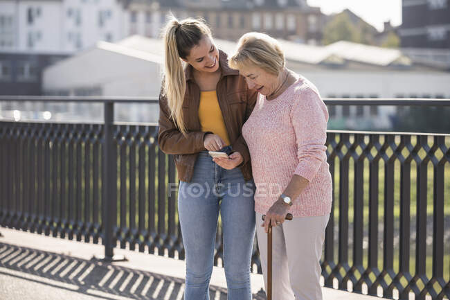 Внучка с бабушкой ходят по пешеходному мосту и пользуются смартфоном — стоковое фото