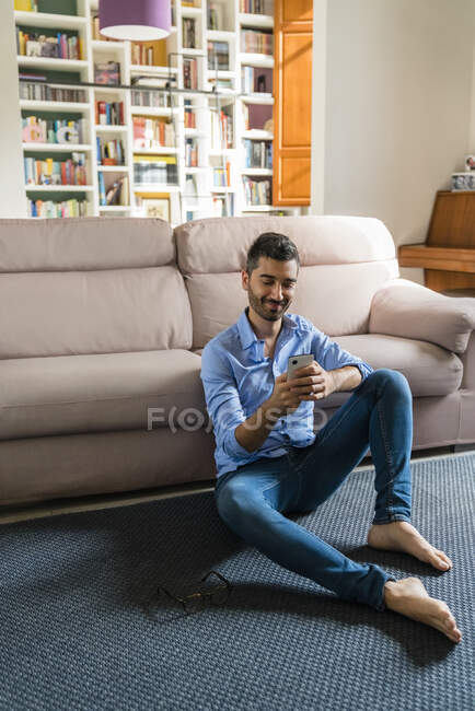 Portrait of smiling young man sitting on the floor of living room at home using smartphone — Stock Photo