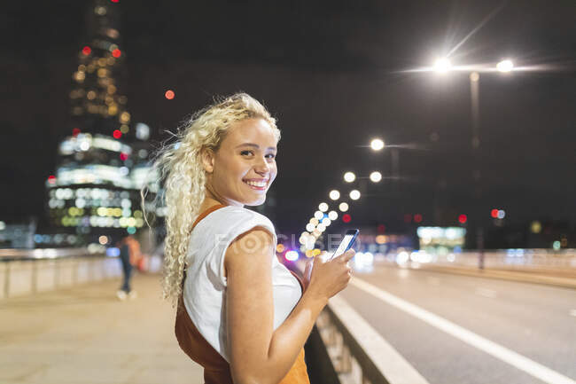 Mujer joven en Londres por la noche y usando su teléfono inteligente - foto de stock