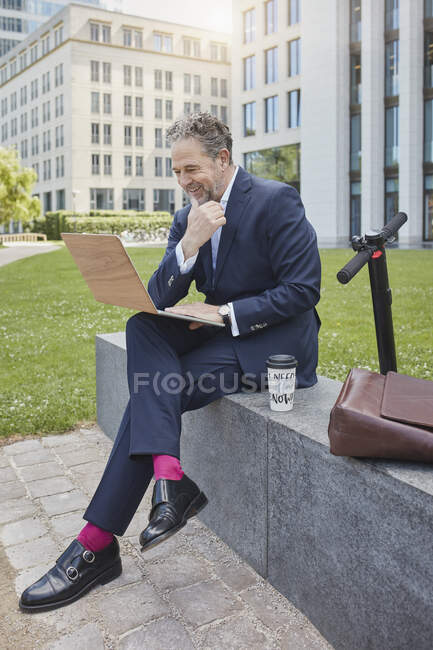 Empresário maduro feliz sentado em uma parede na cidade usando laptop — Fotografia de Stock