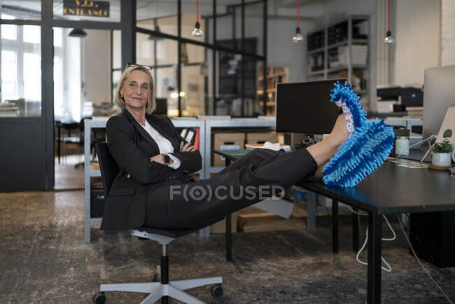 Femme d'affaires mature avec les pieds sur le bureau portant des pantoufles nettoyantes au bureau — Photo de stock