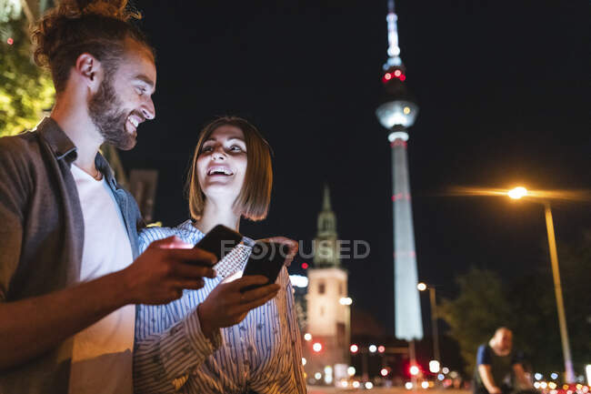 Happy couple using smartphones in the city at night, Fernsehturm in the background, Berlin, Germany — Stock Photo