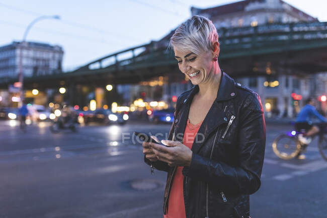 Retrato de la mujer rubia riendo de pie al borde de la carretera por la noche usando el teléfono móvil, Berlín, Alemania - foto de stock